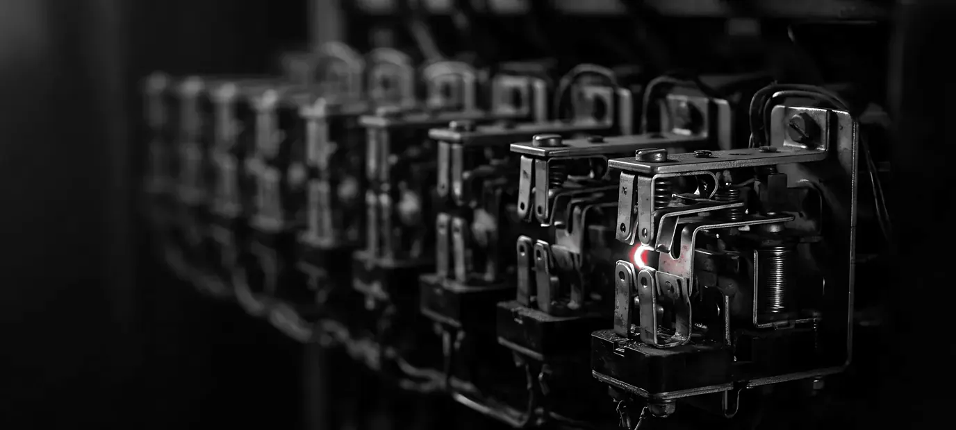 A row of telephone-exchange relays in the dark; only the nearest one has closed, with a small red spark between its contacts